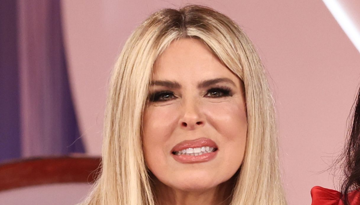Donna con occhi lucidi risponde a domande in uno studio televisivo, mani intrecciate sul tavolo
